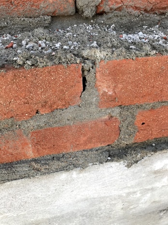 A close-up view of a brick wall segment featuring red bricks and concrete mortar. A noticeable crack runs vertically through one of the bricks, revealing rough and uneven edges. The mortar appears somewhat loose and crumbly in areas.