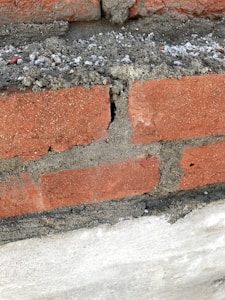 A close-up view of a brick wall segment featuring red bricks and concrete mortar. A noticeable crack runs vertically through one of the bricks, revealing rough and uneven edges. The mortar appears somewhat loose and crumbly in areas.