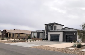 Modern suburban houses with clean lines and a minimalist design are visible. The first house has a multi-car garage and is painted in neutral colors with dark accents. The landscaping includes rocks and drought-resistant plants.