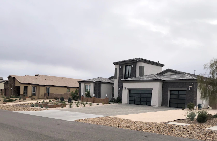 Modern suburban houses with clean lines and a minimalist design are visible. The first house has a multi-car garage and is painted in neutral colors with dark accents. The landscaping includes rocks and drought-resistant plants.