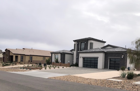 Modern suburban houses with clean lines and a minimalist design are visible. The first house has a multi-car garage and is painted in neutral colors with dark accents. The landscaping includes rocks and drought-resistant plants.