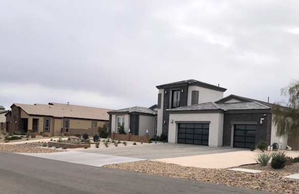 Modern suburban houses with clean lines and a minimalist design are visible. The first house has a multi-car garage and is painted in neutral colors with dark accents. The landscaping includes rocks and drought-resistant plants.