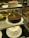 A display of three cakes in a glass case: a chocolate cake with a slice missing on the middle shelf, a white frosted cake with a smooth finish on the top shelf, and a uniquely shaped white frosted cake with pointed peaks adorned with small gold leaf accents on the bottom shelf. Each cake is presented on a white plate.