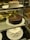 A display of three cakes in a glass case: a chocolate cake with a slice missing on the middle shelf, a white frosted cake with a smooth finish on the top shelf, and a uniquely shaped white frosted cake with pointed peaks adorned with small gold leaf accents on the bottom shelf. Each cake is presented on a white plate.