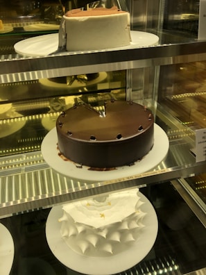 A display of three cakes in a glass case: a chocolate cake with a slice missing on the middle shelf, a white frosted cake with a smooth finish on the top shelf, and a uniquely shaped white frosted cake with pointed peaks adorned with small gold leaf accents on the bottom shelf. Each cake is presented on a white plate.