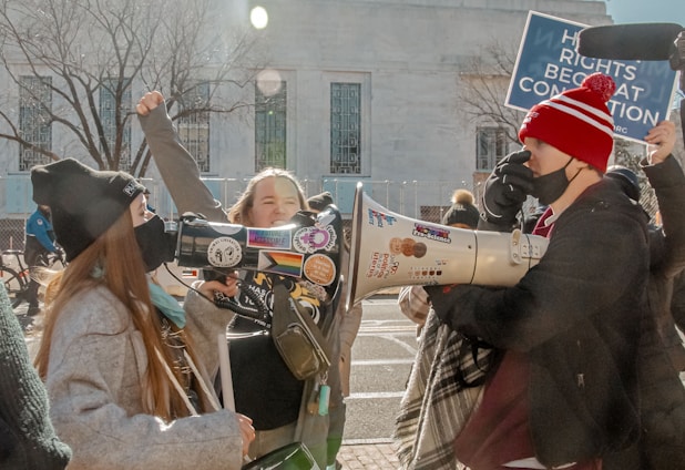 A group of people in a street or plaza setting, wearing winter clothing, and holding megaphones and signs. They appear to be engaged in a protest or demonstration. Stickers on the megaphones feature various slogans and symbols. One person is raising a fist, intensifying the scene's message.