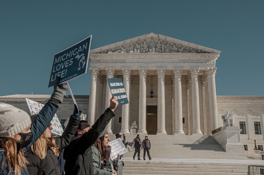 A group of people hold signs in front of a large neoclassical building with tall columns and a detailed pediment. The signs express support for life and human rights. The scene is set on a sunny day, with several people dressed warmly in coats and hats.