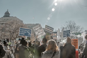 A protest scene with a large group of people holding various signs and banners. The signs display messages related to pro-life and human rights advocacy. The crowd is gathered in front of a government-like building with a dome on top. The sun is shining brightly, casting lens flares across the image, and bare trees suggest a winter setting.