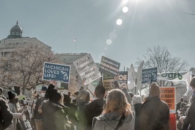 A protest scene with a large group of people holding various signs and banners. The signs display messages related to pro-life and human rights advocacy. The crowd is gathered in front of a government-like building with a dome on top. The sun is shining brightly, casting lens flares across the image, and bare trees suggest a winter setting.