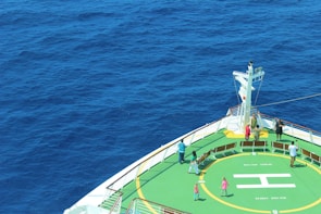 The bow of a ship is visible, featuring a helipad marked with an 'H' and surrounded by a green circular area. Several people are walking or standing around the helipad. The vast expanse of deep blue ocean surrounds the ship, creating a tranquil and serene backdrop.