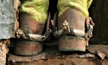 A pair of worn cowboy boots with bright green decorative stitching is seen from the back. The boots have metal spurs attached, indicating they are used for horseback riding. They rest on a rough, rusted metal or wooden surface, suggesting a rugged, outdoor environment.