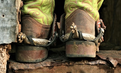 A pair of worn cowboy boots with bright green decorative stitching is seen from the back. The boots have metal spurs attached, indicating they are used for horseback riding. They rest on a rough, rusted metal or wooden surface, suggesting a rugged, outdoor environment.