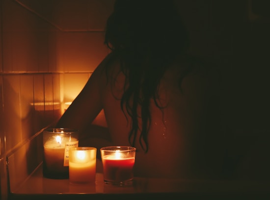A dimly lit scene with a person sitting in a bathtub surrounded by softly glowing candles. The ambiance suggests a calm and intimate setting with focus on relaxation and tranquility.