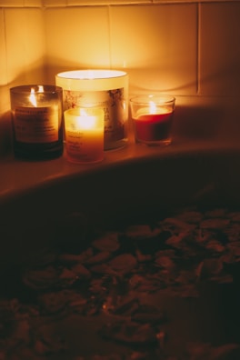 Three lit candles are placed on the edge of a bathtub, casting a warm, soft glow. The surface of the water in the bathtub is covered with scattered rose petals, creating a relaxing atmosphere.
