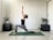 A person is performing a lunge yoga pose on a green yoga mat in a minimalist room. The individual is dressed in a black sleeveless top and patterned leggings, with arms raised and palms together. The room is decorated with a few potted plants placed on dark brown furniture, and there is soft natural light coming through.