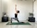 A person is performing a lunge yoga pose on a green yoga mat in a minimalist room. The individual is dressed in a black sleeveless top and patterned leggings, with arms raised and palms together. The room is decorated with a few potted plants placed on dark brown furniture, and there is soft natural light coming through.