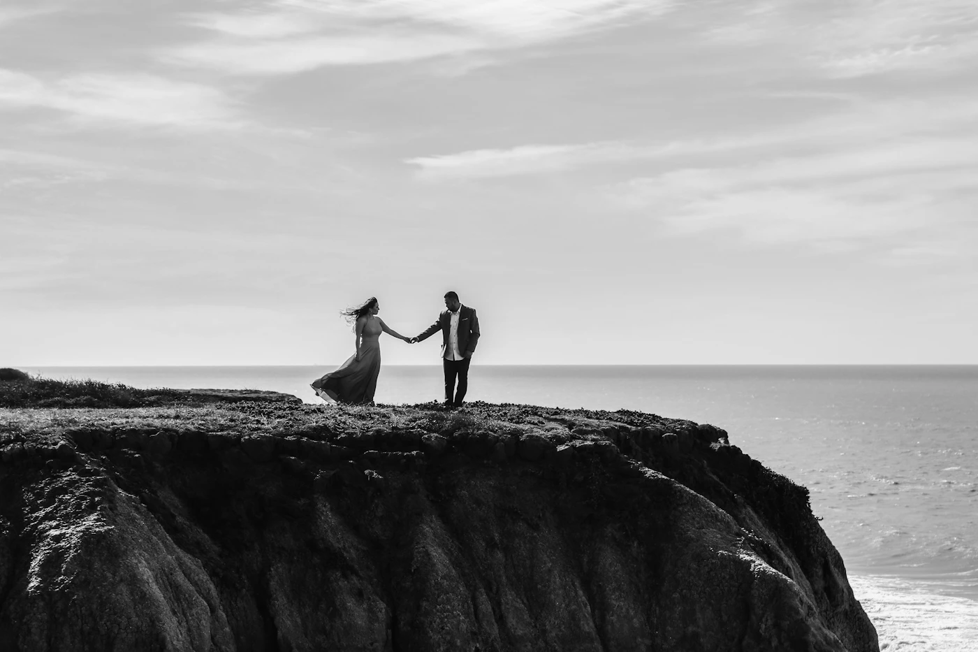 Couple in formal wedding attire standing together on a dramatic coastal cliff with vast ocean stretching to the horizon below