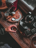 A cozy coffee-making scene with two espresso machines on a wooden surface. A hand holds a small cup of espresso, while several empty cups and saucers are placed nearby. The setting is warmly lit, creating a rustic and inviting atmosphere.