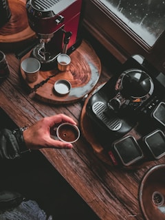 A cozy coffee-making scene with two espresso machines on a wooden surface. A hand holds a small cup of espresso, while several empty cups and saucers are placed nearby. The setting is warmly lit, creating a rustic and inviting atmosphere.