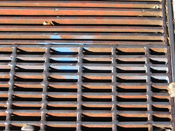 A close-up of a metal grate with vertical and horizontal bars forming a grid pattern. There is orange and rust coloration on the metal along with patches of blue paint. Some leaves and a small stone are visible on and around the grate.