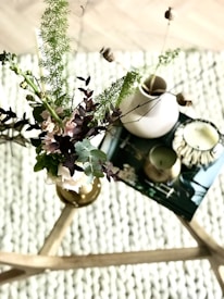 A cozy setting featuring a wooden coffee table adorned with a decorative arrangement of green and purple foliage in a vase. Beside it, there are two candles, one in a metallic holder and another in a ceramic dish, placed on top of a book or magazine. The background reveals a textured, woven surface.
