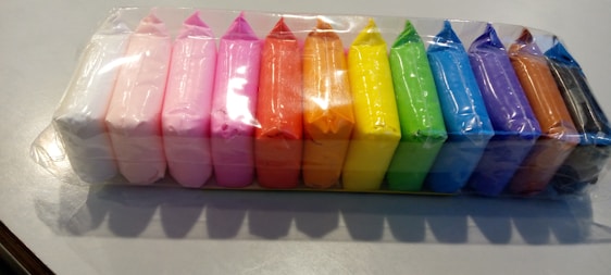 A row of colorful, rectangular clay blocks wrapped in clear plastic packaging. The blocks include a variety of colors such as white, pink, red, orange, yellow, green, blue, purple, brown, and black. They are neatly arranged in a single line on a light gray surface.
