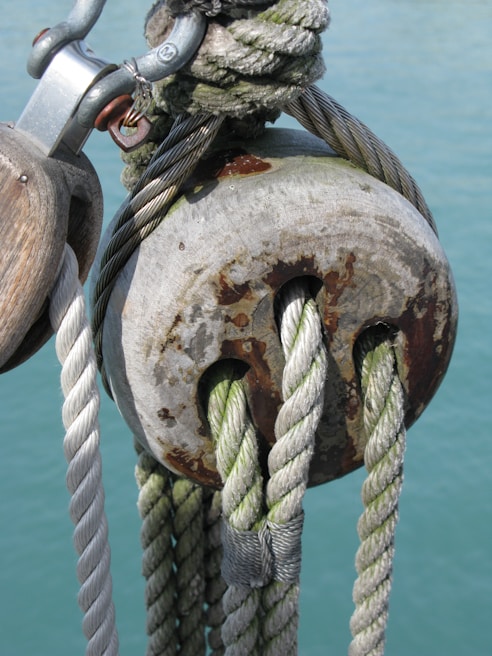 Weathered nautical rope and metal pulley showcase signs of use and exposure to the elements. The ropes are intertwined through a metallic fitting, with rust visible on the metal surfaces. The background is a calm, teal body of water, enhancing the maritime theme.