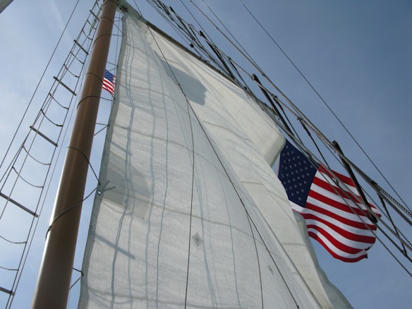 A sailboat's mast with several large white sails unfurled, billowing against a backdrop of clear blue sky. An American flag is prominently displayed, waving in the breeze. The rigging and lines are visible, adding a sense of detail and structure to the image.
