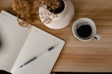 A notebook with lined pages labeled 'Reflection' lies open on a wooden table, with a pen resting on its pages. Nearby, there is a white ceramic mug filled with black coffee. Some dried flowers in a white vase are also present on the table.