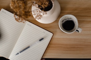 A notebook with lined pages labeled 'Reflection' lies open on a wooden table, with a pen resting on its pages. Nearby, there is a white ceramic mug filled with black coffee. Some dried flowers in a white vase are also present on the table.
