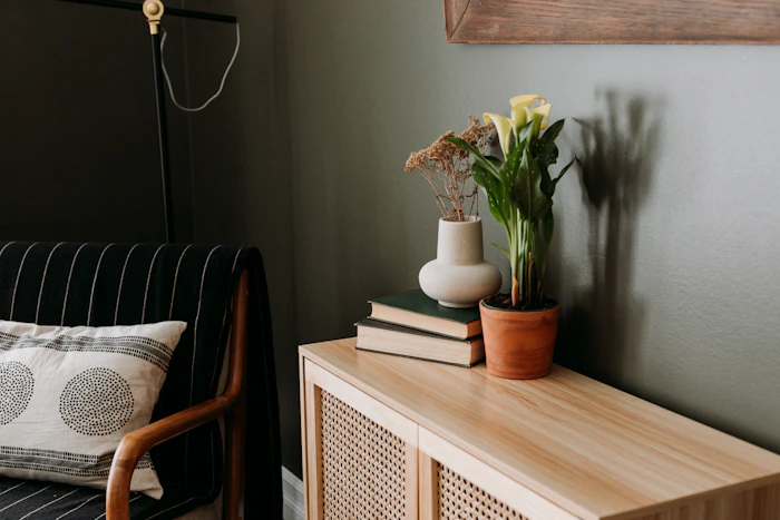 Corner with an olive wall, a rattan cabinet, stacked books and a vase of yellow flowers