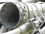 A close-up view of industrial machinery, featuring metal pipes, valves, and a large cylindrical component. The equipment is composed of various metallic surfaces, with visible screws and clamps securing the parts together. The texture of the metal appears weathered.