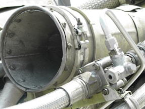 A close-up view of industrial machinery, featuring metal pipes, valves, and a large cylindrical component. The equipment is composed of various metallic surfaces, with visible screws and clamps securing the parts together. The texture of the metal appears weathered.