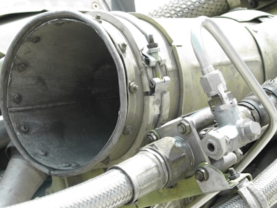 A close-up view of industrial machinery, featuring metal pipes, valves, and a large cylindrical component. The equipment is composed of various metallic surfaces, with visible screws and clamps securing the parts together. The texture of the metal appears weathered.