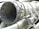 A close-up view of industrial machinery, featuring metal pipes, valves, and a large cylindrical component. The equipment is composed of various metallic surfaces, with visible screws and clamps securing the parts together. The texture of the metal appears weathered.