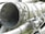 A close-up view of industrial machinery, featuring metal pipes, valves, and a large cylindrical component. The equipment is composed of various metallic surfaces, with visible screws and clamps securing the parts together. The texture of the metal appears weathered.
