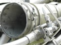 A close-up view of industrial machinery, featuring metal pipes, valves, and a large cylindrical component. The equipment is composed of various metallic surfaces, with visible screws and clamps securing the parts together. The texture of the metal appears weathered.