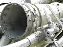 A close-up view of industrial machinery, featuring metal pipes, valves, and a large cylindrical component. The equipment is composed of various metallic surfaces, with visible screws and clamps securing the parts together. The texture of the metal appears weathered.