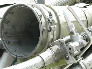 A close-up view of industrial machinery, featuring metal pipes, valves, and a large cylindrical component. The equipment is composed of various metallic surfaces, with visible screws and clamps securing the parts together. The texture of the metal appears weathered.
