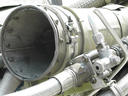 A close-up view of industrial machinery, featuring metal pipes, valves, and a large cylindrical component. The equipment is composed of various metallic surfaces, with visible screws and clamps securing the parts together. The texture of the metal appears weathered.