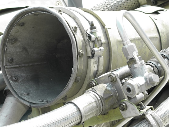 A close-up view of industrial machinery, featuring metal pipes, valves, and a large cylindrical component. The equipment is composed of various metallic surfaces, with visible screws and clamps securing the parts together. The texture of the metal appears weathered.
