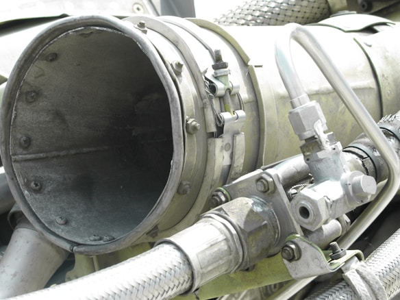 A close-up view of industrial machinery, featuring metal pipes, valves, and a large cylindrical component. The equipment is composed of various metallic surfaces, with visible screws and clamps securing the parts together. The texture of the metal appears weathered.