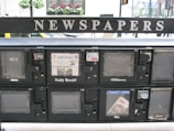 A series of newspaper vending machines lined up along a street. Each machine displays various newspapers, including the 'Daily Herald' and 'Hoy,' with the words 'NEWSPAPERS' prominently displayed above the machines. Some newspapers are behind protective glass, and the background features a busy street scene with cars and pedestrians.