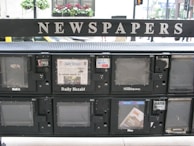 A series of newspaper vending machines lined up along a street. Each machine displays various newspapers, including the 'Daily Herald' and 'Hoy,' with the words 'NEWSPAPERS' prominently displayed above the machines. Some newspapers are behind protective glass, and the background features a busy street scene with cars and pedestrians.