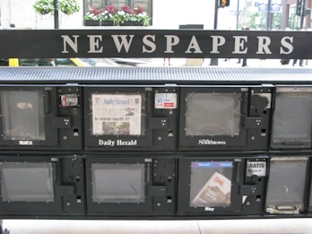 A series of newspaper vending machines lined up along a street. Each machine displays various newspapers, including the 'Daily Herald' and 'Hoy,' with the words 'NEWSPAPERS' prominently displayed above the machines. Some newspapers are behind protective glass, and the background features a busy street scene with cars and pedestrians.