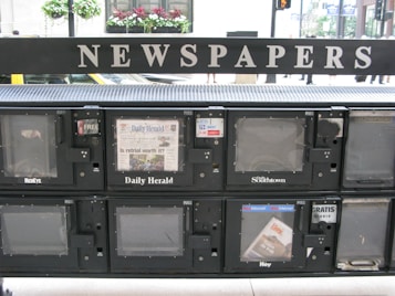 A series of newspaper vending machines lined up along a street. Each machine displays various newspapers, including the 'Daily Herald' and 'Hoy,' with the words 'NEWSPAPERS' prominently displayed above the machines. Some newspapers are behind protective glass, and the background features a busy street scene with cars and pedestrians.