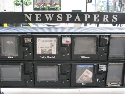 A series of newspaper vending machines lined up along a street. Each machine displays various newspapers, including the 'Daily Herald' and 'Hoy,' with the words 'NEWSPAPERS' prominently displayed above the machines. Some newspapers are behind protective glass, and the background features a busy street scene with cars and pedestrians.