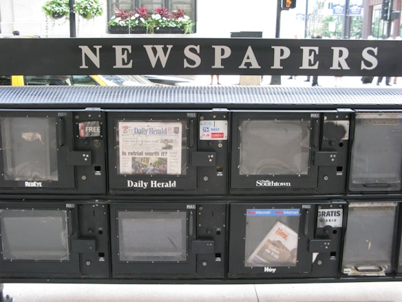 A series of newspaper vending machines lined up along a street. Each machine displays various newspapers, including the 'Daily Herald' and 'Hoy,' with the words 'NEWSPAPERS' prominently displayed above the machines. Some newspapers are behind protective glass, and the background features a busy street scene with cars and pedestrians.