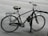 A bicycle is securely locked to a black metal bike rack on the side of a paved street. The bicycle has a black seat with noticeable wear and tear and a rear rack. The background shows an empty road with no visible traffic.