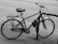 A bicycle is securely locked to a black metal bike rack on the side of a paved street. The bicycle has a black seat with noticeable wear and tear and a rear rack. The background shows an empty road with no visible traffic.